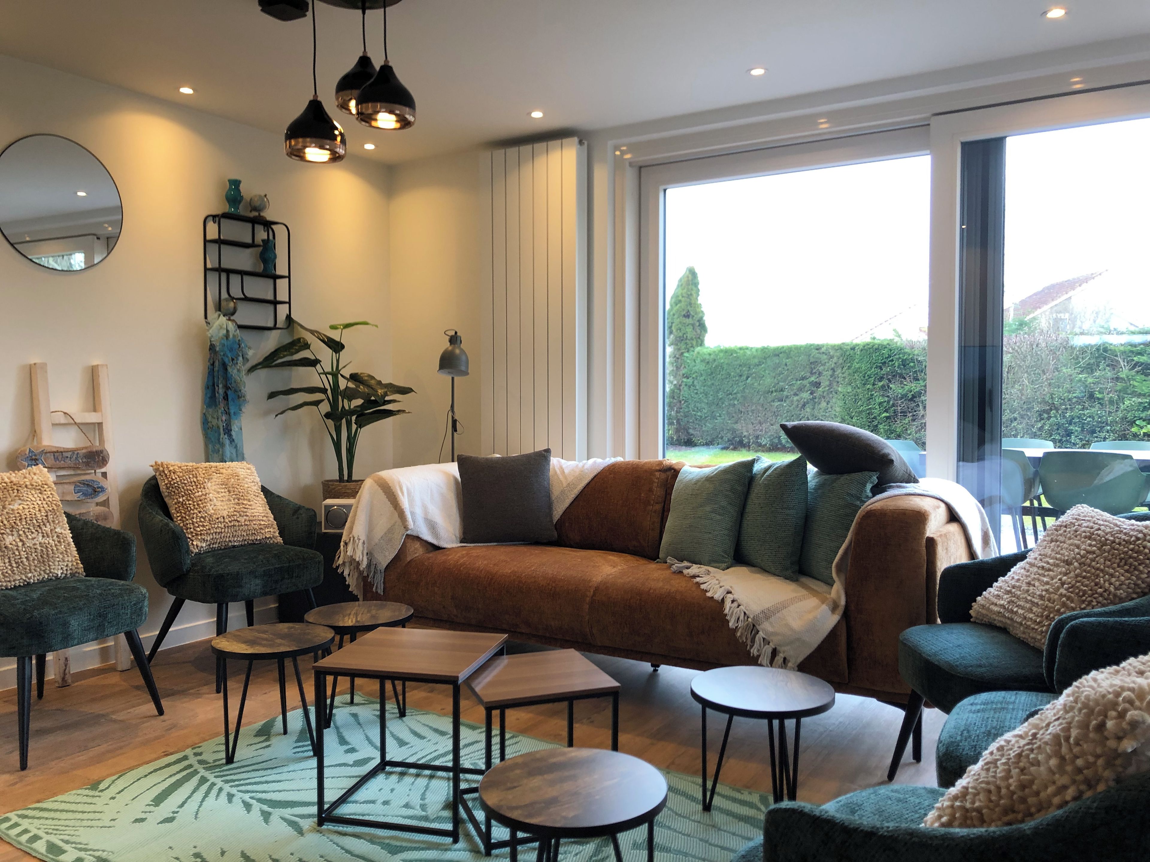 Een moderne woonkamer met een bank, fauteuils en kleine tafels. Een groot raam met uitzicht op groen.