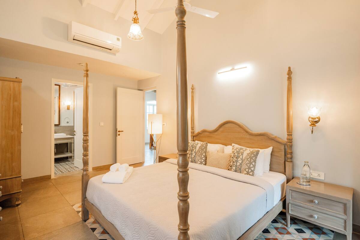 A four-poster bed with a wooden headboard, a bedside table with a lamp, and a bathroom visible through an open door.