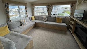A living room with a grey sofa, a television, and a view of mobile homes. - Greenacres Holiday Park - Holiday Accommodation 15761 (Porthmadog)
