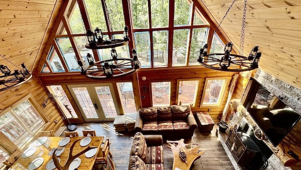 A log cabin interior with a high ceiling, large windows, a fireplace, a dining table with antler chairs, and a sofa. - A Custom-Built Swiss-Style Luxury Mountain Retreat - Upper Level 3 BR Unit (Mount Jackson)