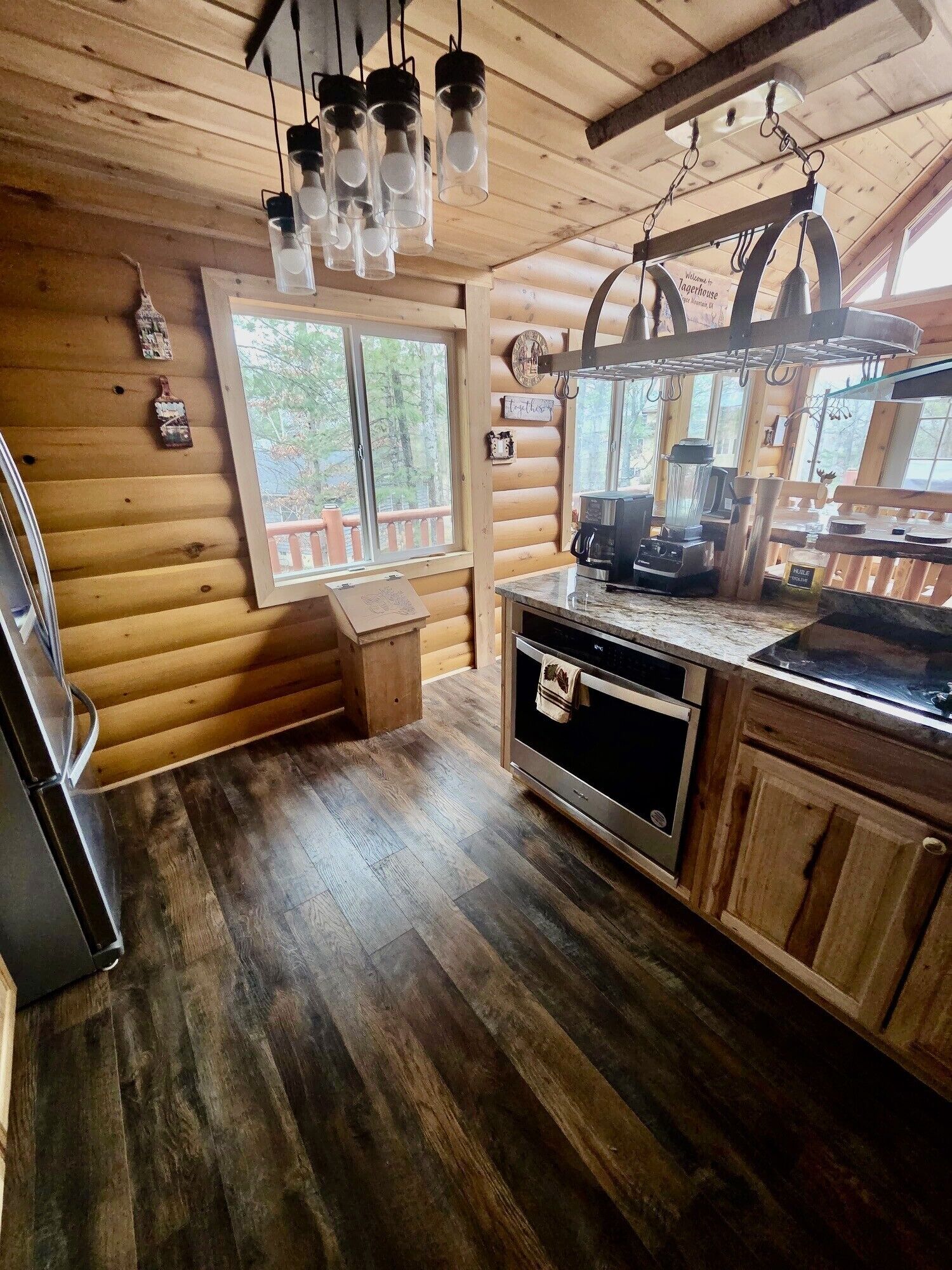 A log cabin kitchen with wooden walls, a wooden floor, a window, a sink, a stove, and hanging light fixtures.