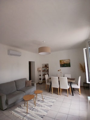 Ein Wohnzimmer mit Sofa, Couchtisch, Esstisch mit Stühlen, einer Lampe und einem Bild an der Wand.