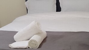 Cama bem arrumada com lençóis brancos e duas toalhas enroladas, cabeceira escura e parede lisa.