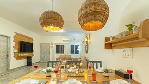 Ein Essbereich mit einem Holztisch, gedeckt für vier Personen, umgeben von Korbschwinglampen, einem an der Wand befestigten Fernseher und einer Couch im Hintergrund.