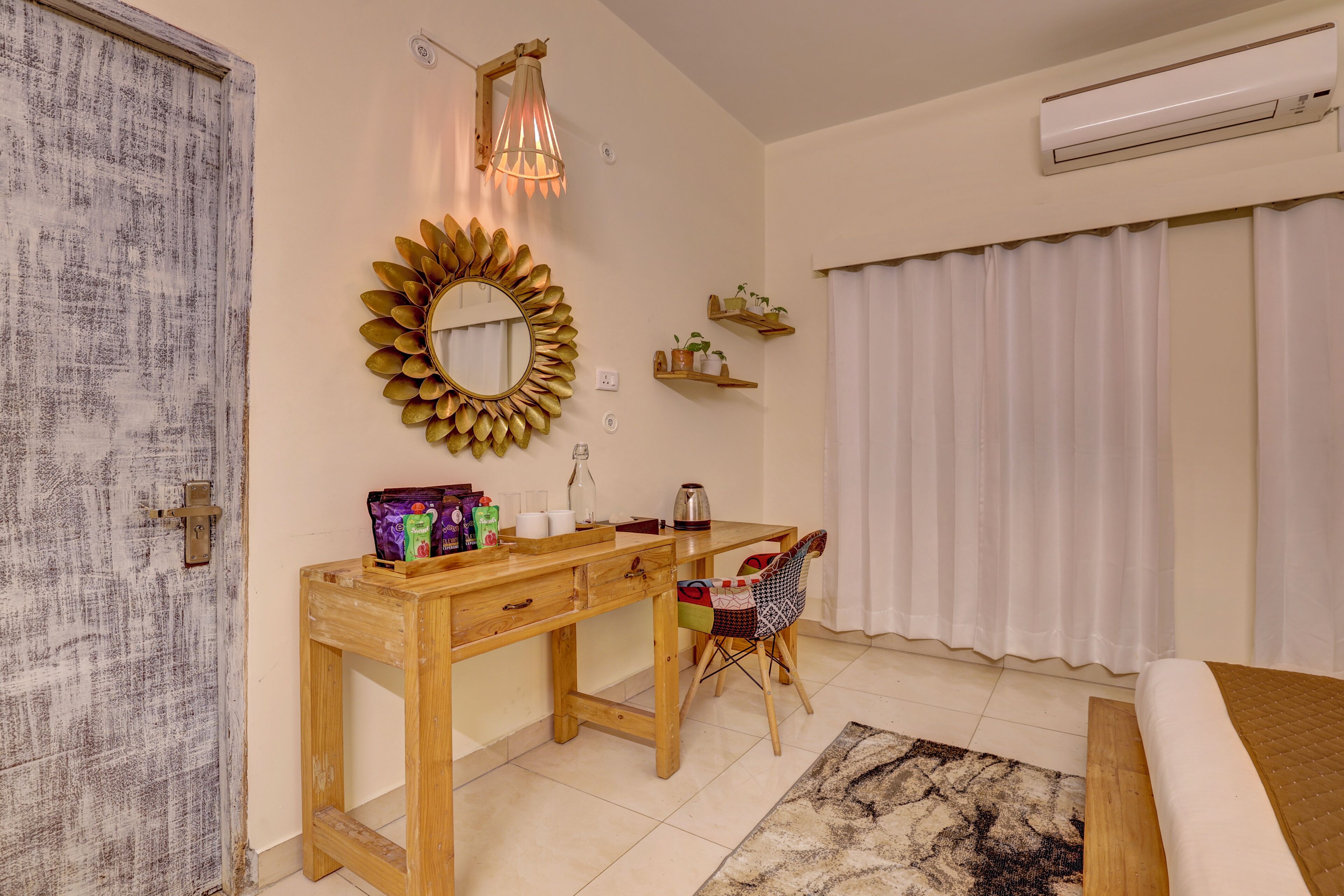 A bedroom with a wooden desk, a chair, a bed, a mirror with a sunflower design, a hanging light fixture, and a window with curtains.