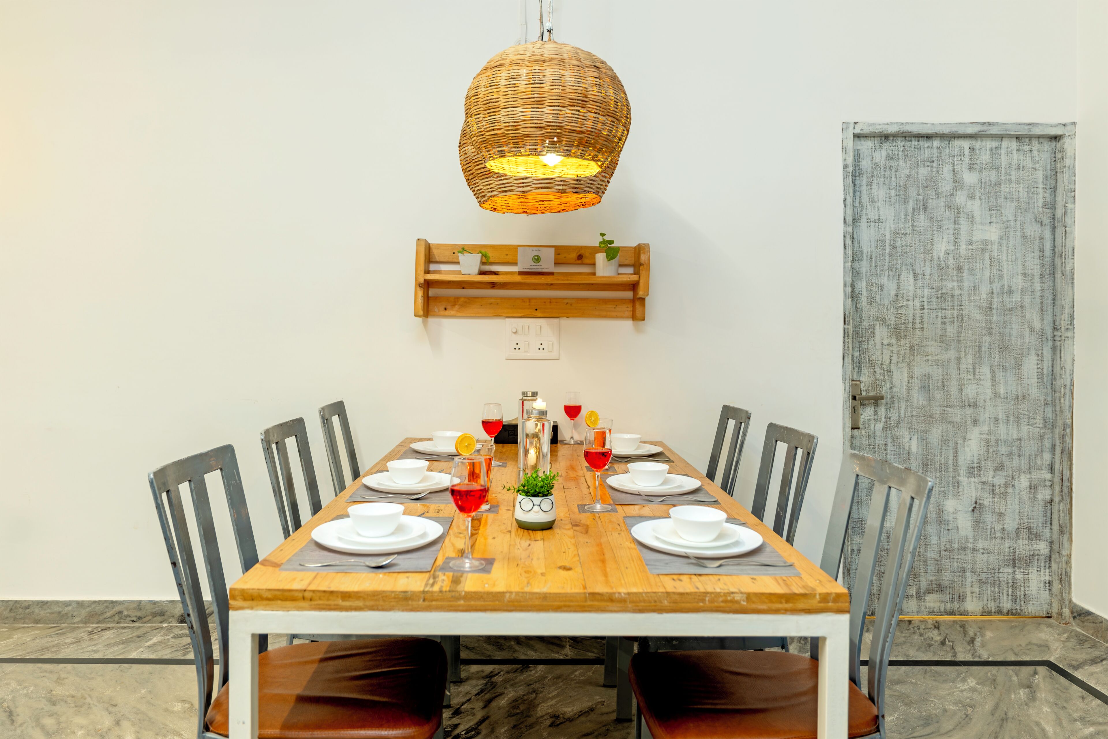 A dining table with chairs, a hanging light fixture, a wall-mounted shelf with plants, and a door.
