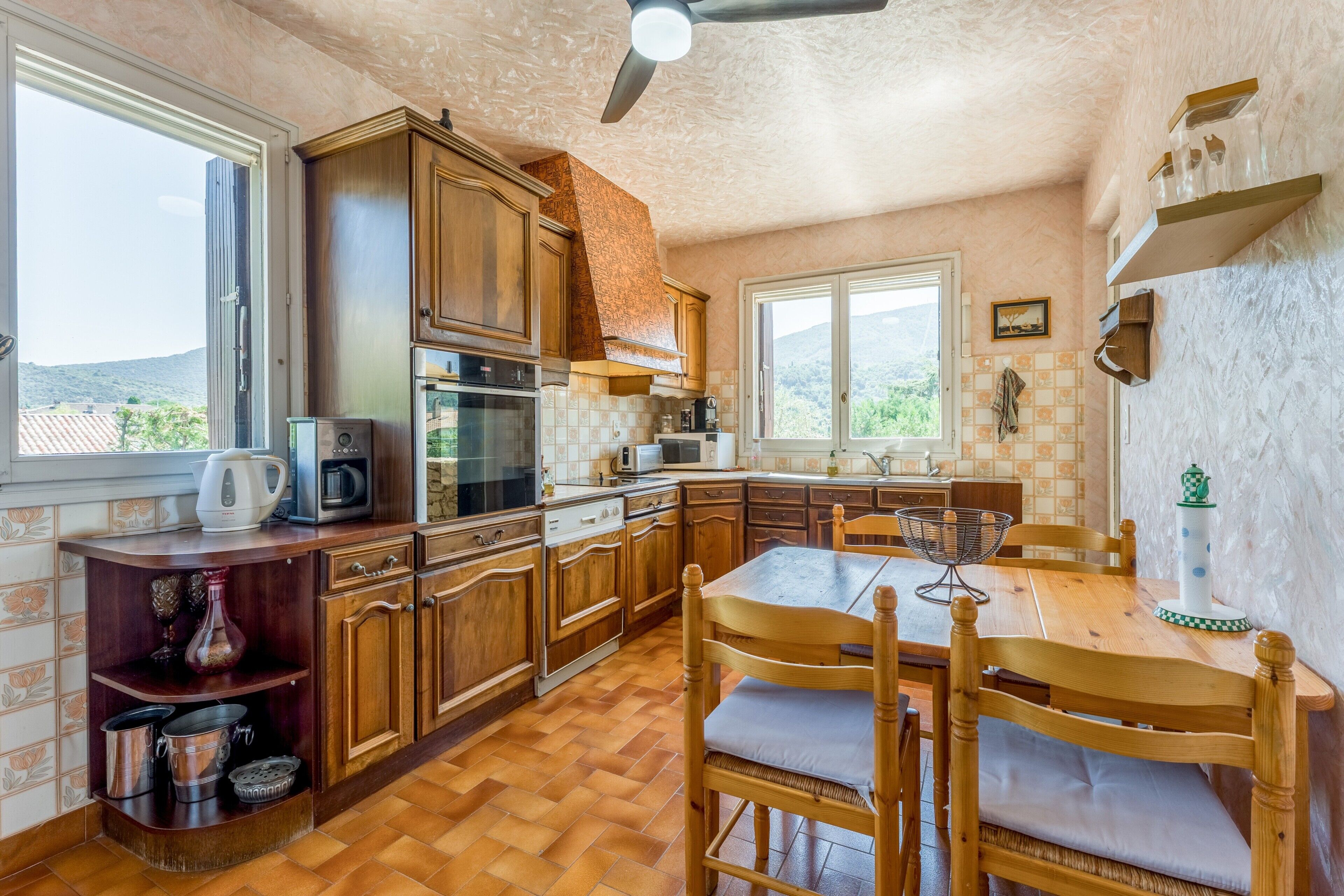 A kitchen with wooden cabinets, a dining table with chairs, a ceiling fan, and a window with a view of the outdoors.