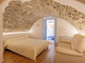 Ein Schlafzimmer mit Steinwand, einem Bett, einem Sessel und Blick durch eine offene Tür.