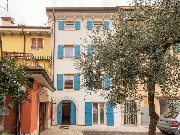 Ein mehrstöckiges Gebäude mit blauen Fensterläden, einem gut sichtbaren Eingang und einem Baum im Vordergrund.