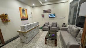 A living room with a marble reception desk, grey sofas, a coffee table, and a TV mounted on the wall. - Hotel Pilgrrim (Varanasi)