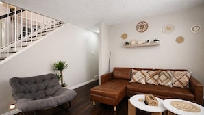 Una sala de estar con un sillón gris tapizado, un sofá de cuero marrón, una mesita blanca y una escalera.