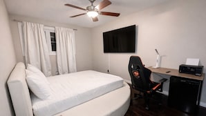 Una habitación pequeña con cama, escritorio, silla, ventilador de techo y televisión.