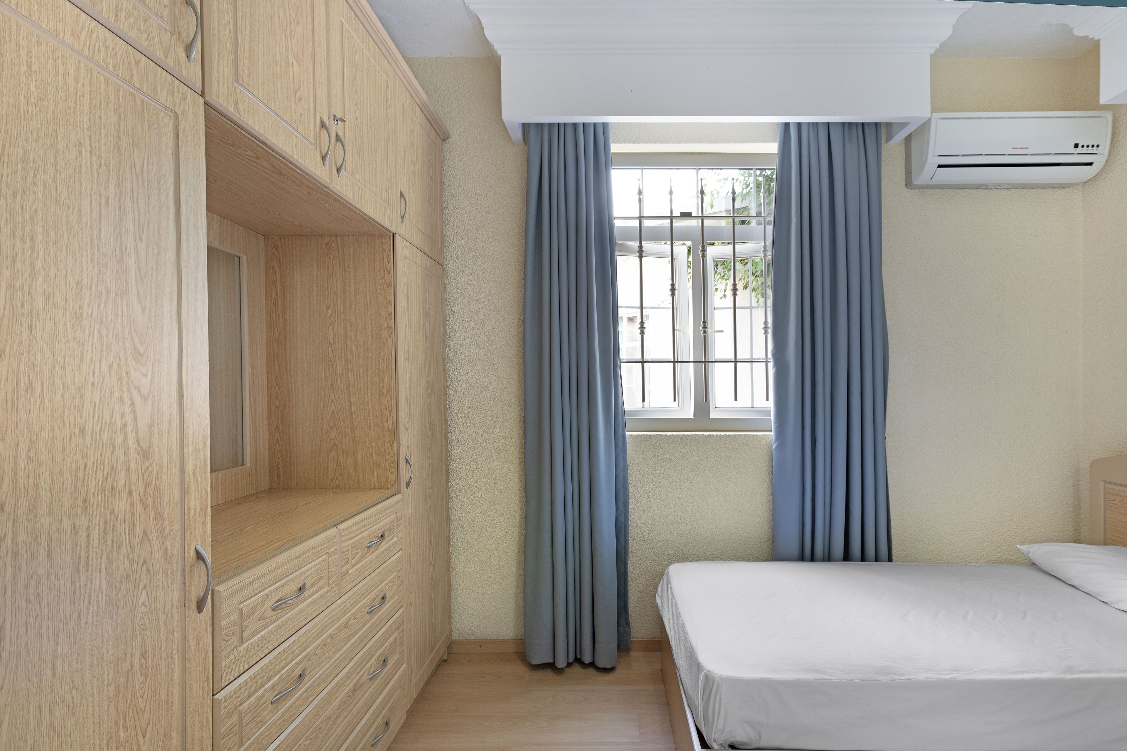 A bedroom with a bed, a wardrobe, a window with curtains, and an air conditioning unit.