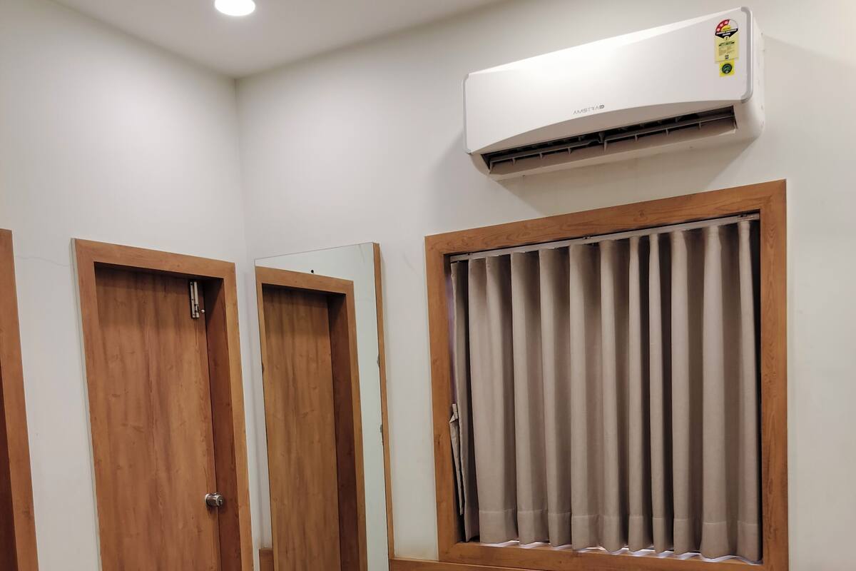 A hotel room with a bed, two wooden doors, an air conditioning unit, a window with curtains, and a wall-mounted light.