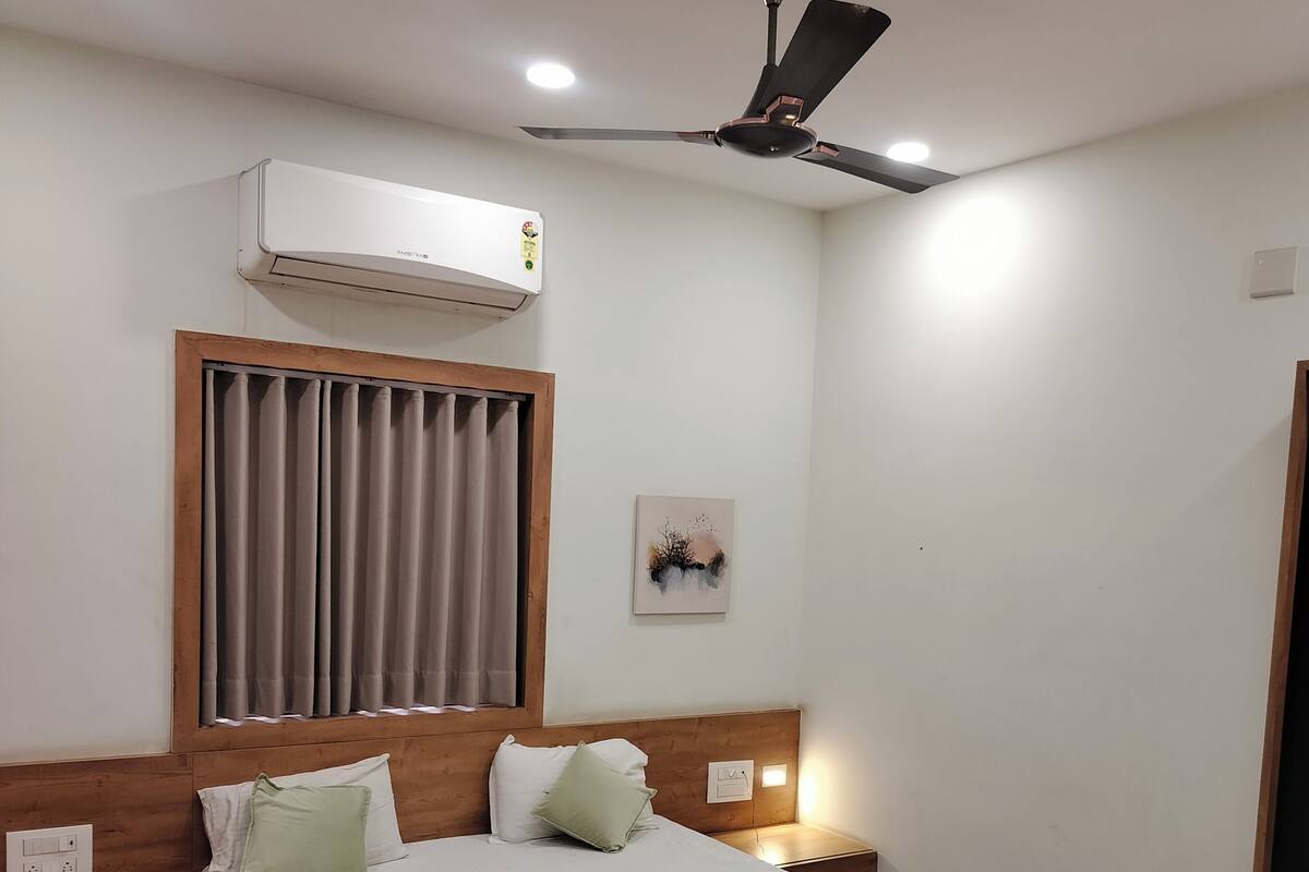 A bedroom with a bed, wooden headboard, a ceiling fan, an air conditioner, a bedside table, and a wall-mounted light.