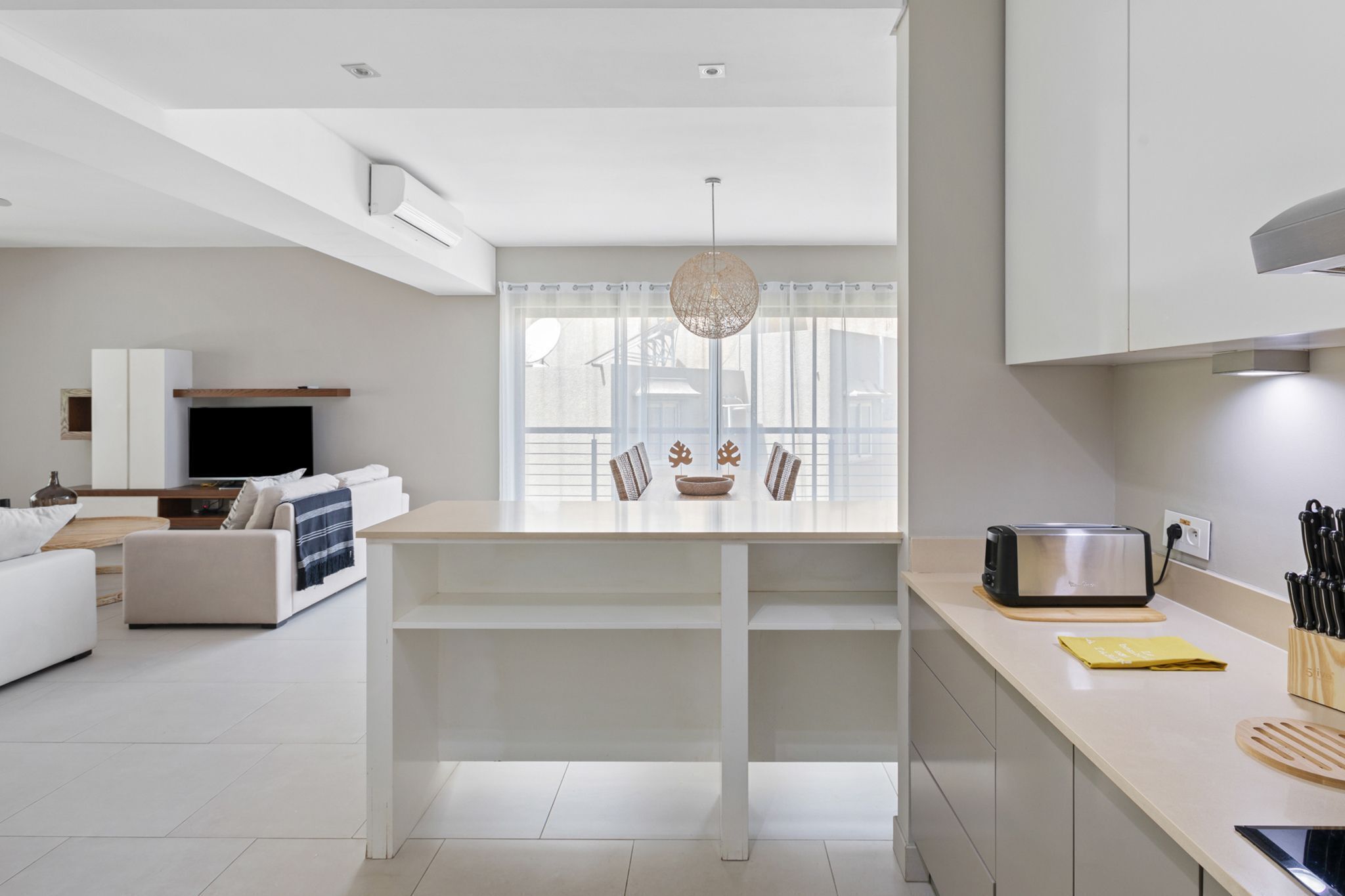 Een moderne keuken met een centraal kookeiland, een eethoek met een witte tafel en een woonkamer met een bank en televisie.