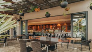 Un espace café en plein air avec des chaises et des tables en osier, des plantes suspendues et une vue sur le bar intérieur.