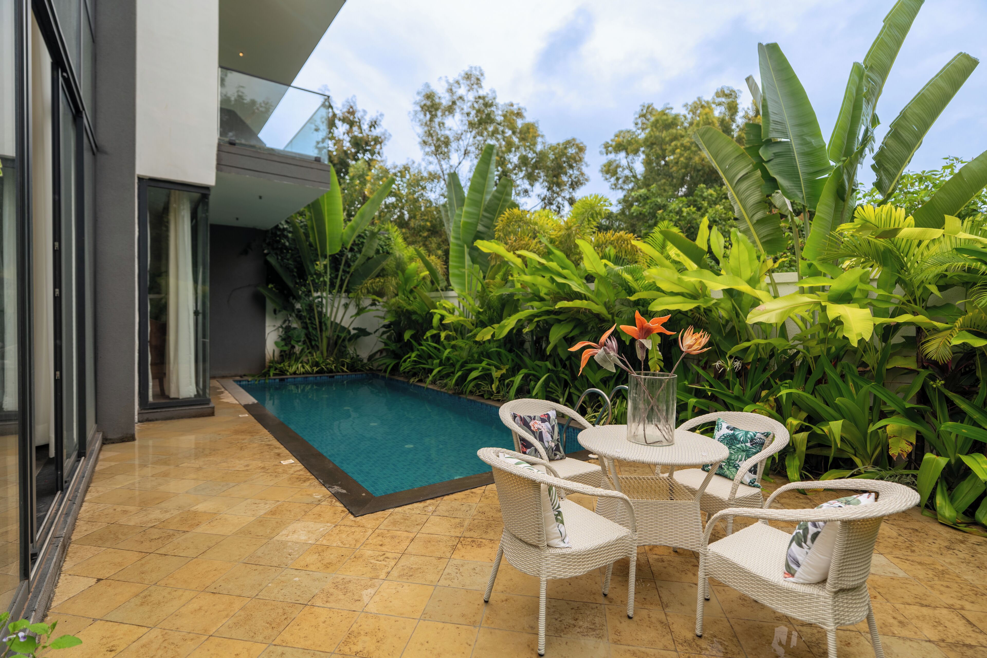 Uma área moderna de piscina ao ar livre com um terraço envidraçado, cercada por exuberantes plantas tropicais e mobiliada com cadeiras e mesa de vime.