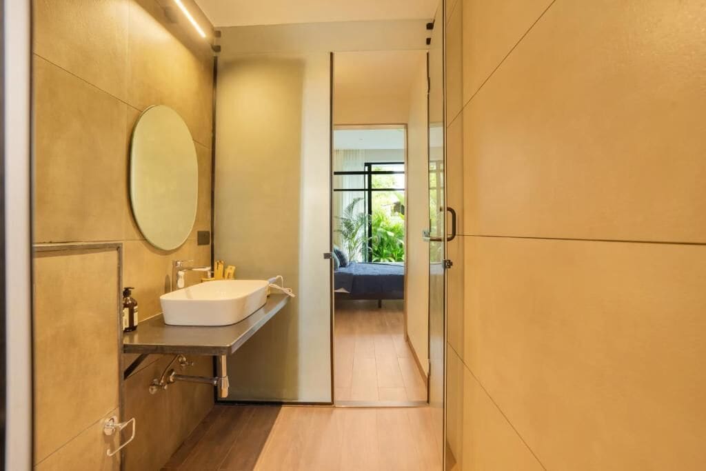 A modern bathroom with a large mirror, a sink, and a glass door leading to a bedroom.