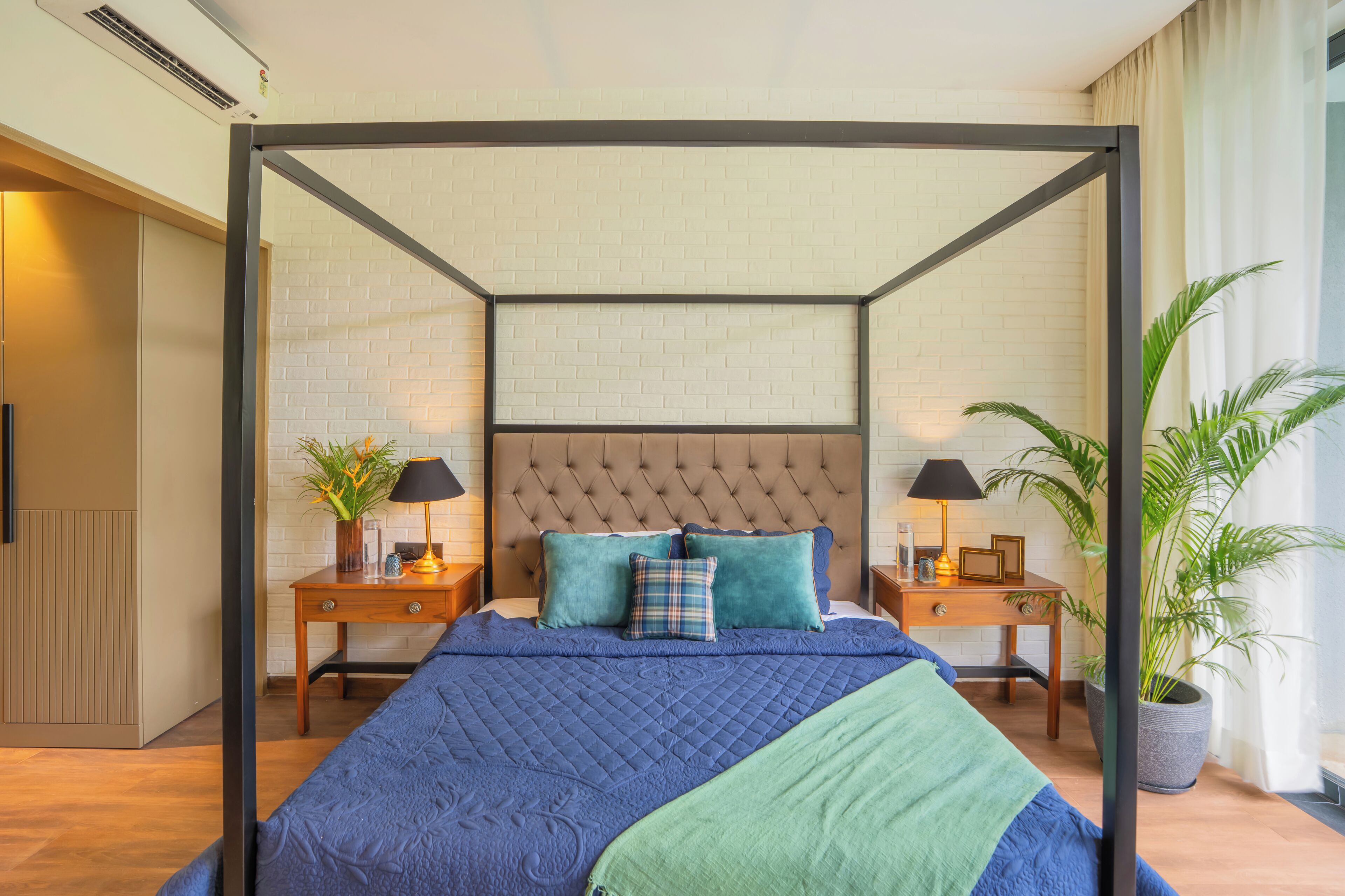A bedroom with a four-poster bed, a wooden nightstand with a lamp, a potted plant, and a view of the outside.