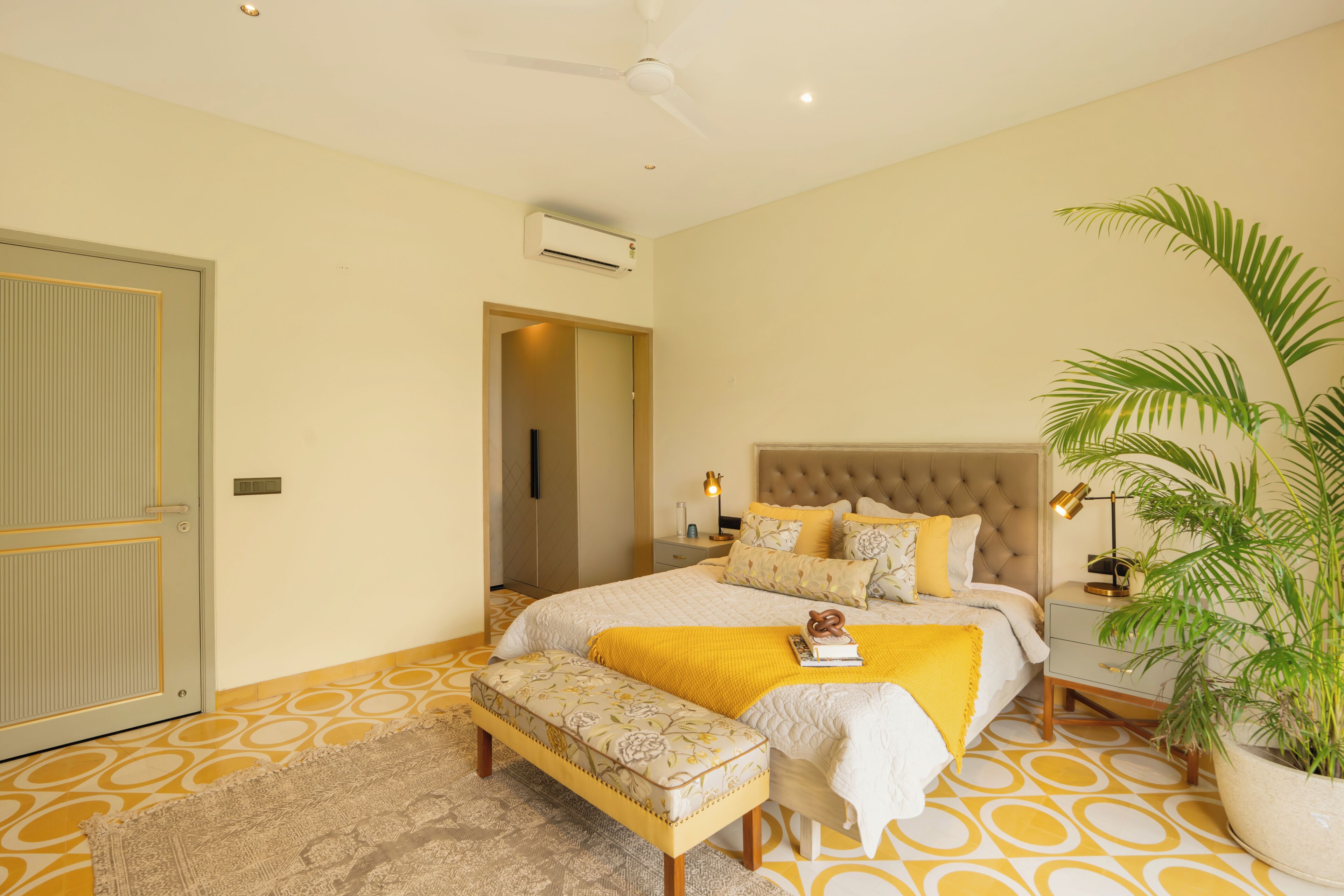 A bedroom with a bed, bedside tables, a bench, a potted plant, and an air conditioning unit.