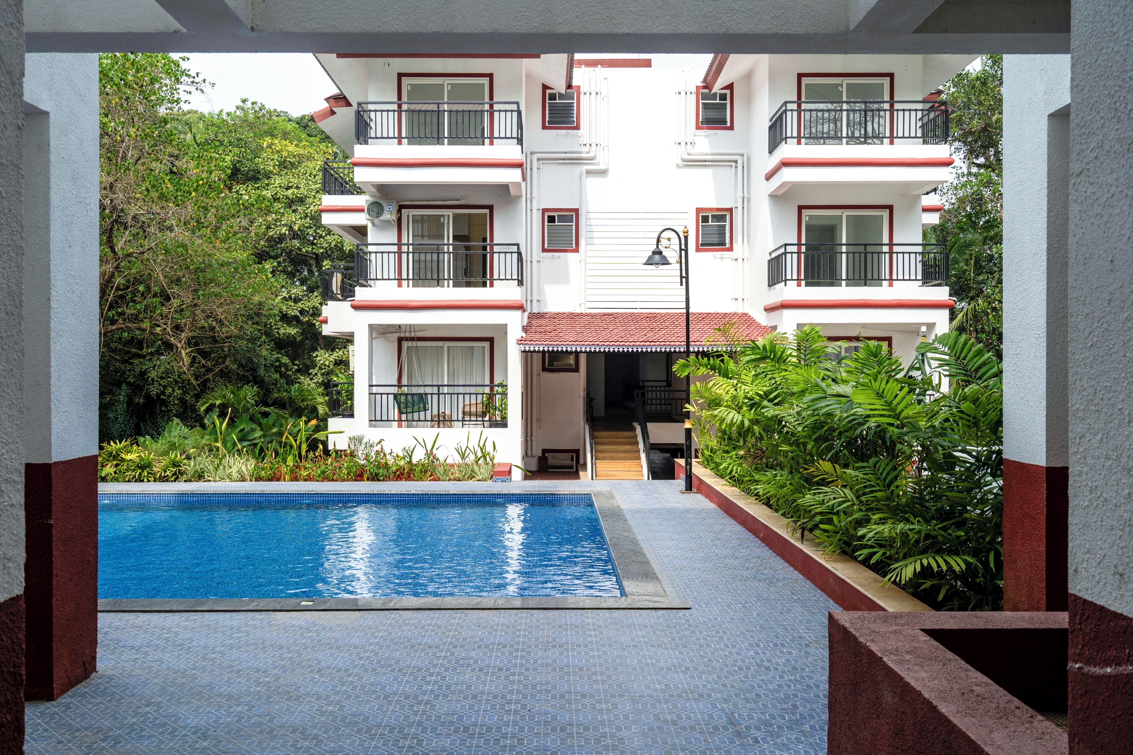 A swimming pool with a tiled deck, surrounded by lush greenery and a multi-story building with balconies.