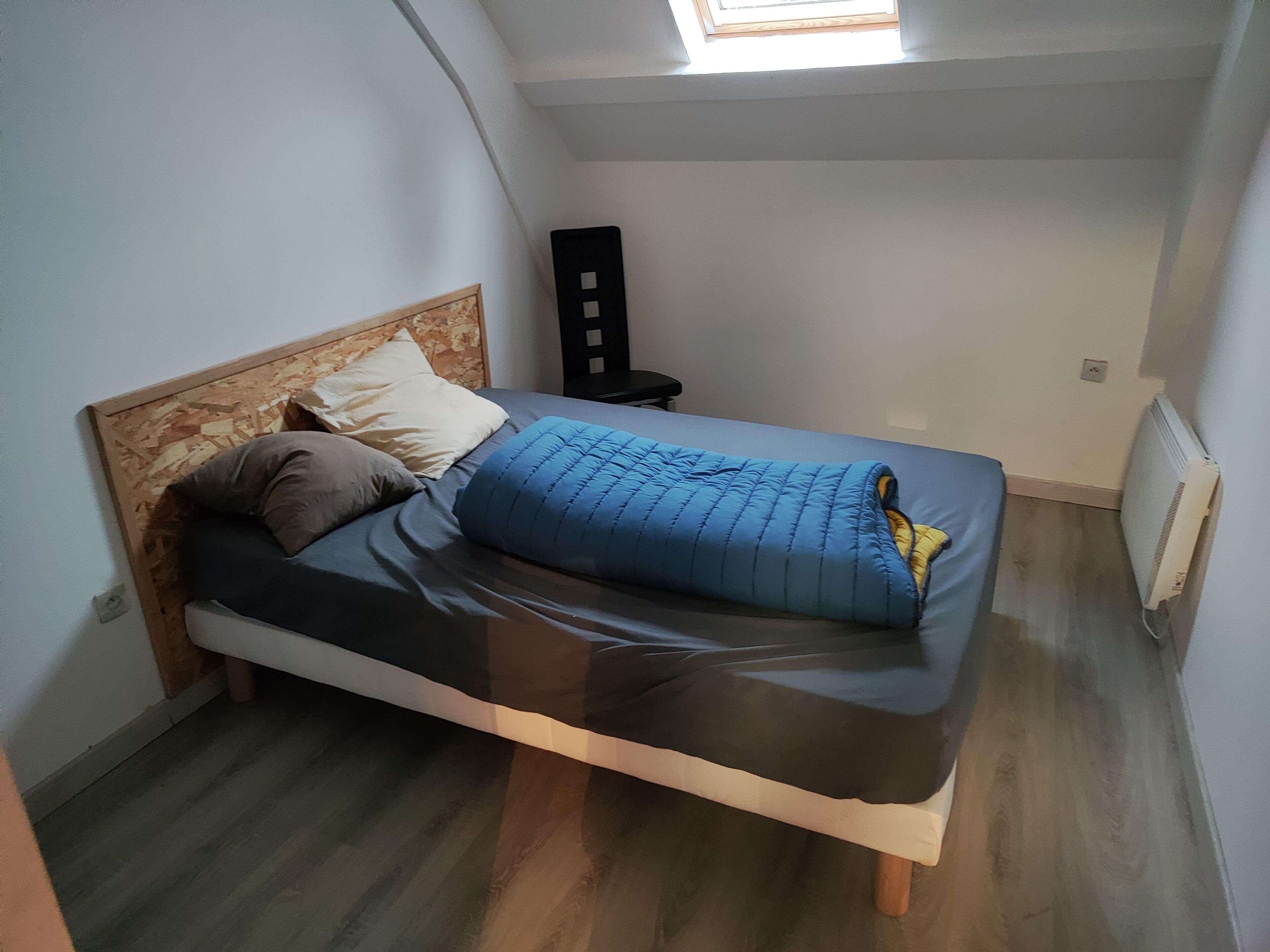 Ein Einzelbett mit Holz-Kopfteil, einer blauen Bettdecke und zwei Kissen. Ein kleiner Nachttisch steht daneben.
