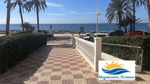 Ein gepflasterter Weg mit Palmen, Blick auf das Meer und geparkten Autos.