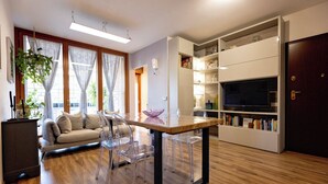 Een moderne woonkamer met een bank, transparante stoelen, een houten tafel en een ingebouwde boekenkast met een televisie.