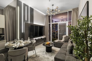 Un salon moderne doté d’un téléviseur à écran plat, d’une table à manger, d’un canapé gris et d’une grande fenêtre offrant une vue sur la ville.