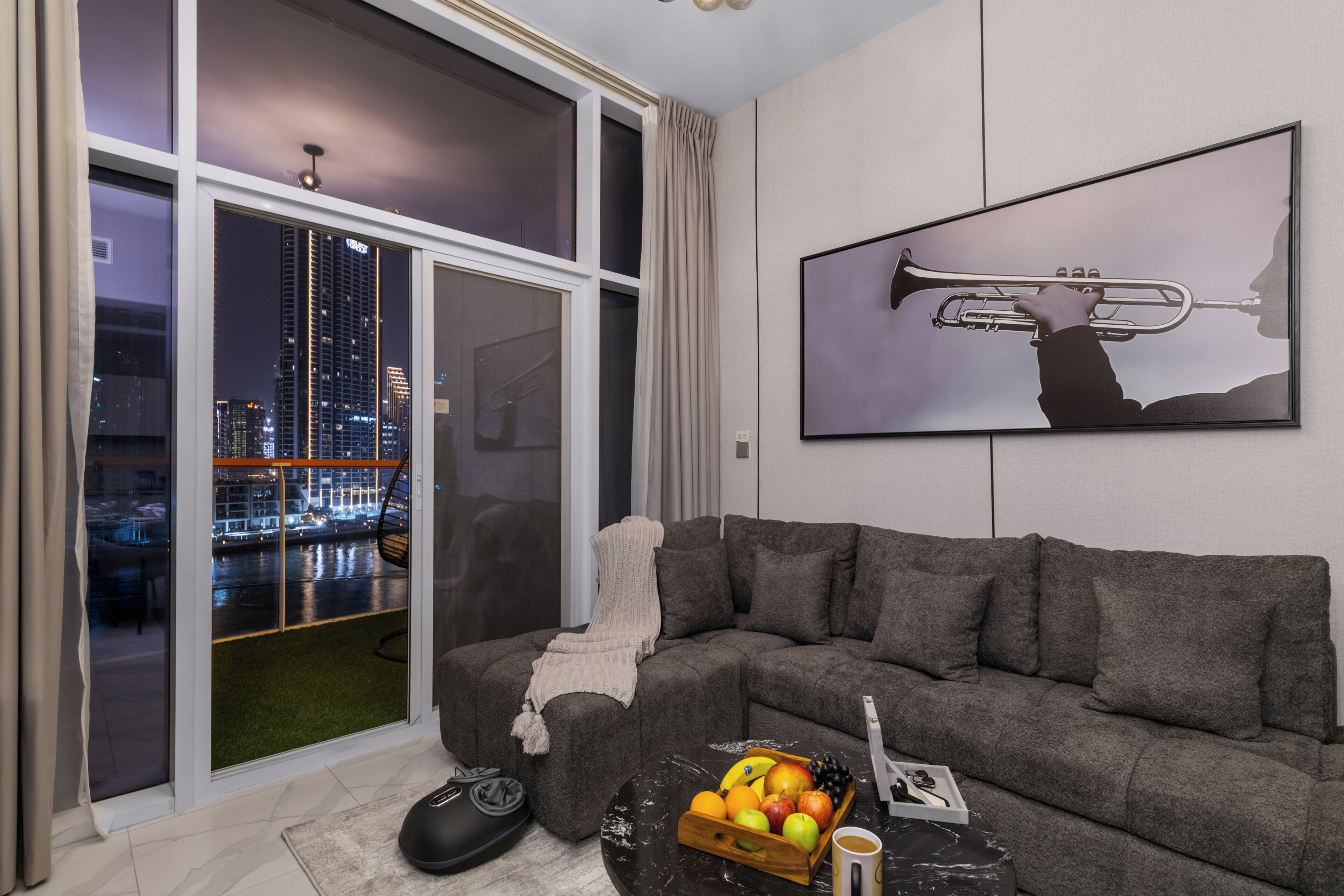Ein modernes Wohnzimmer mit einem Ecksofa, einem großen Fenster mit Blick auf die Stadt, einem aufgehängten Bild einer Trompete und einer Obstschale auf einem Couchtisch.