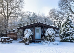 Une cabane en bois dont l’entrée est recouverte de neige, avec un banc et un arbre également recouvert de neige au premier plan.