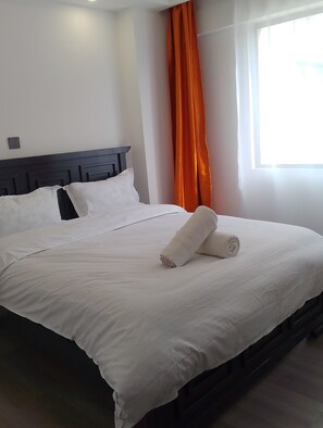 A hotel room with a neatly made bed, a dark headboard, an orange curtain, and a window with natural light. - Cozy 1-bedroom apartment in serene Nairobi with fitness room Lavington-Kilimani (Nairobi)