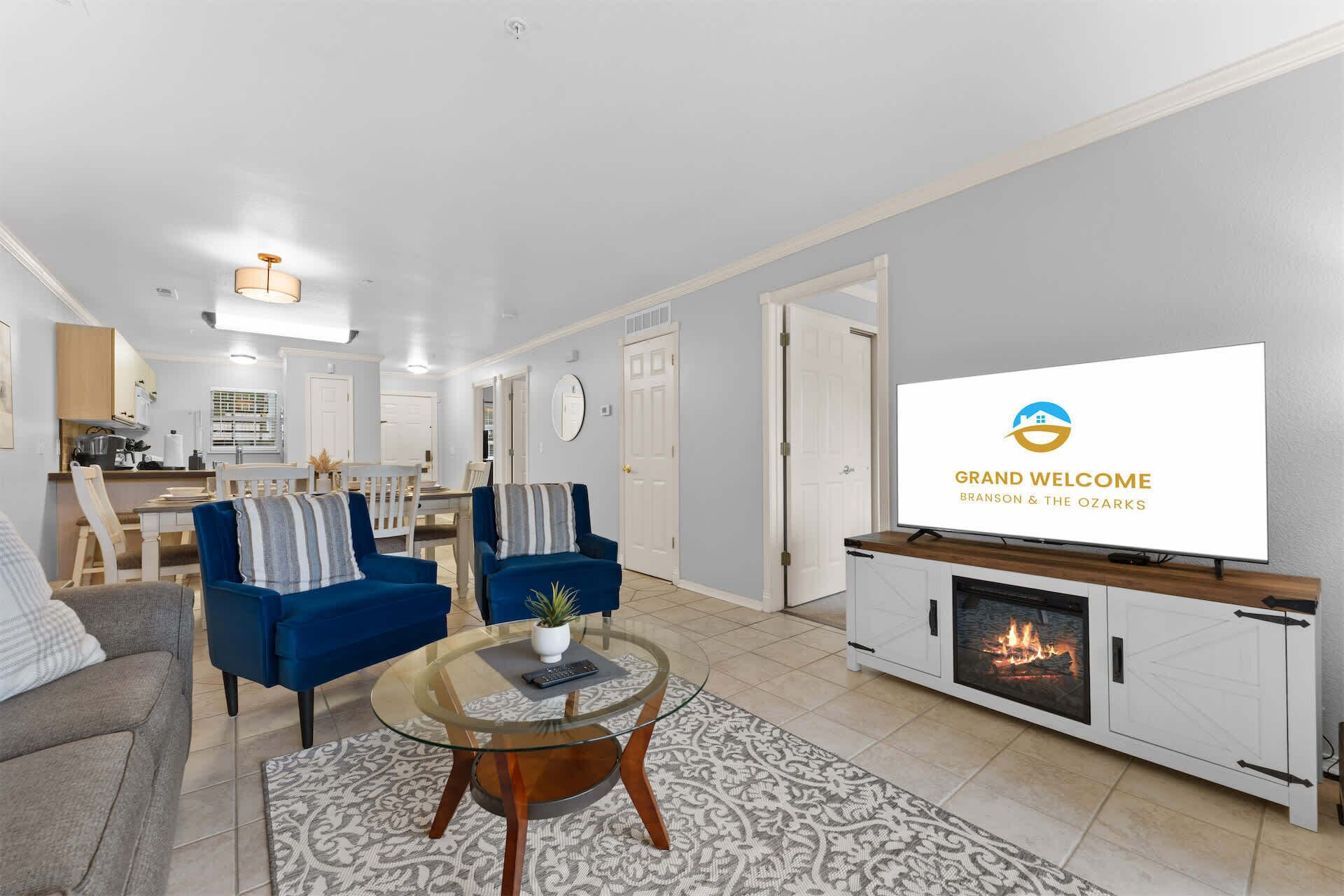 A modern living room with a fireplace, a television displaying a welcome message, and a dining area in the background.