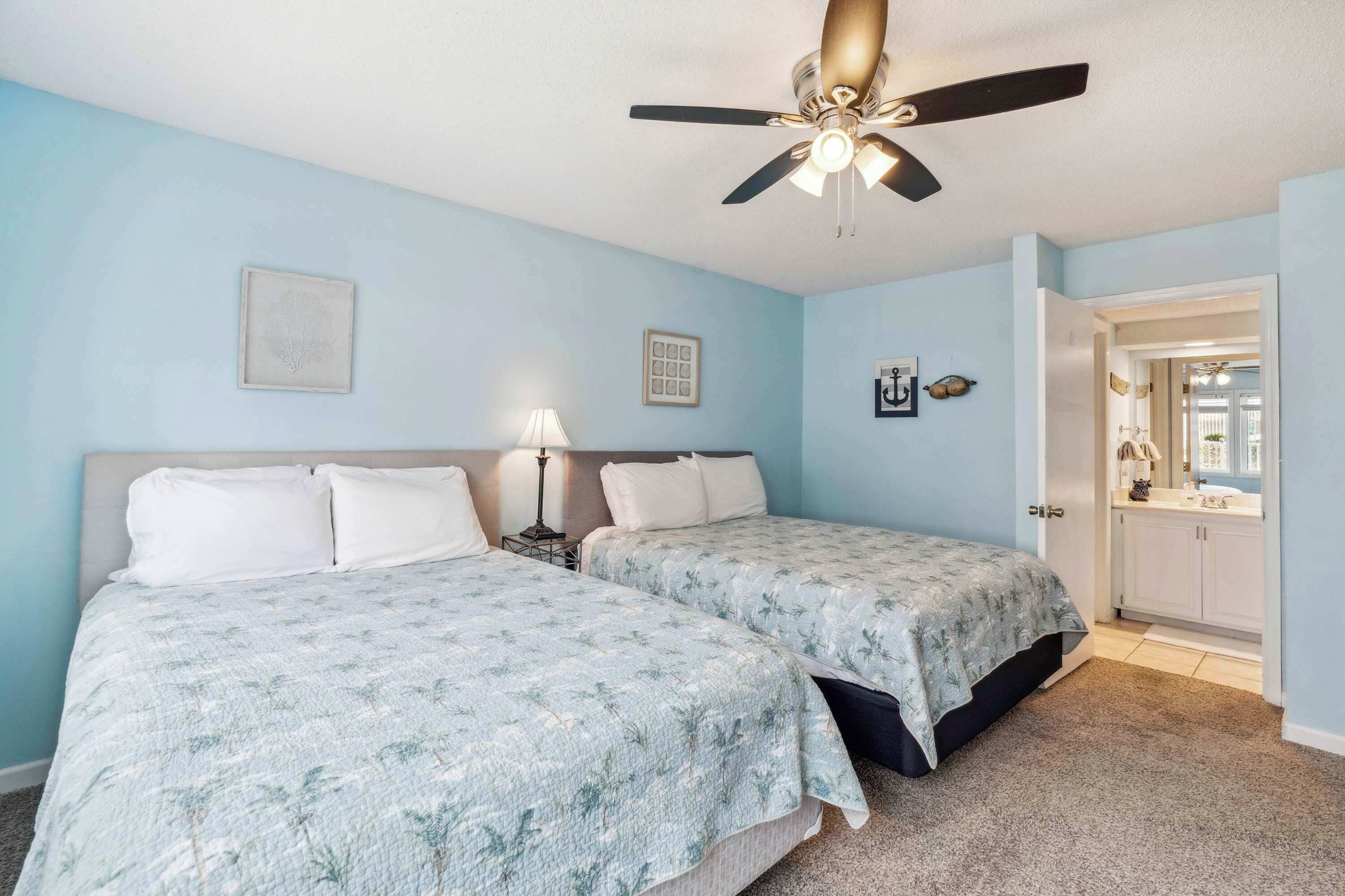 A bedroom with two beds, a ceiling fan, a nightstand with a lamp, and a bathroom visible through an open door.