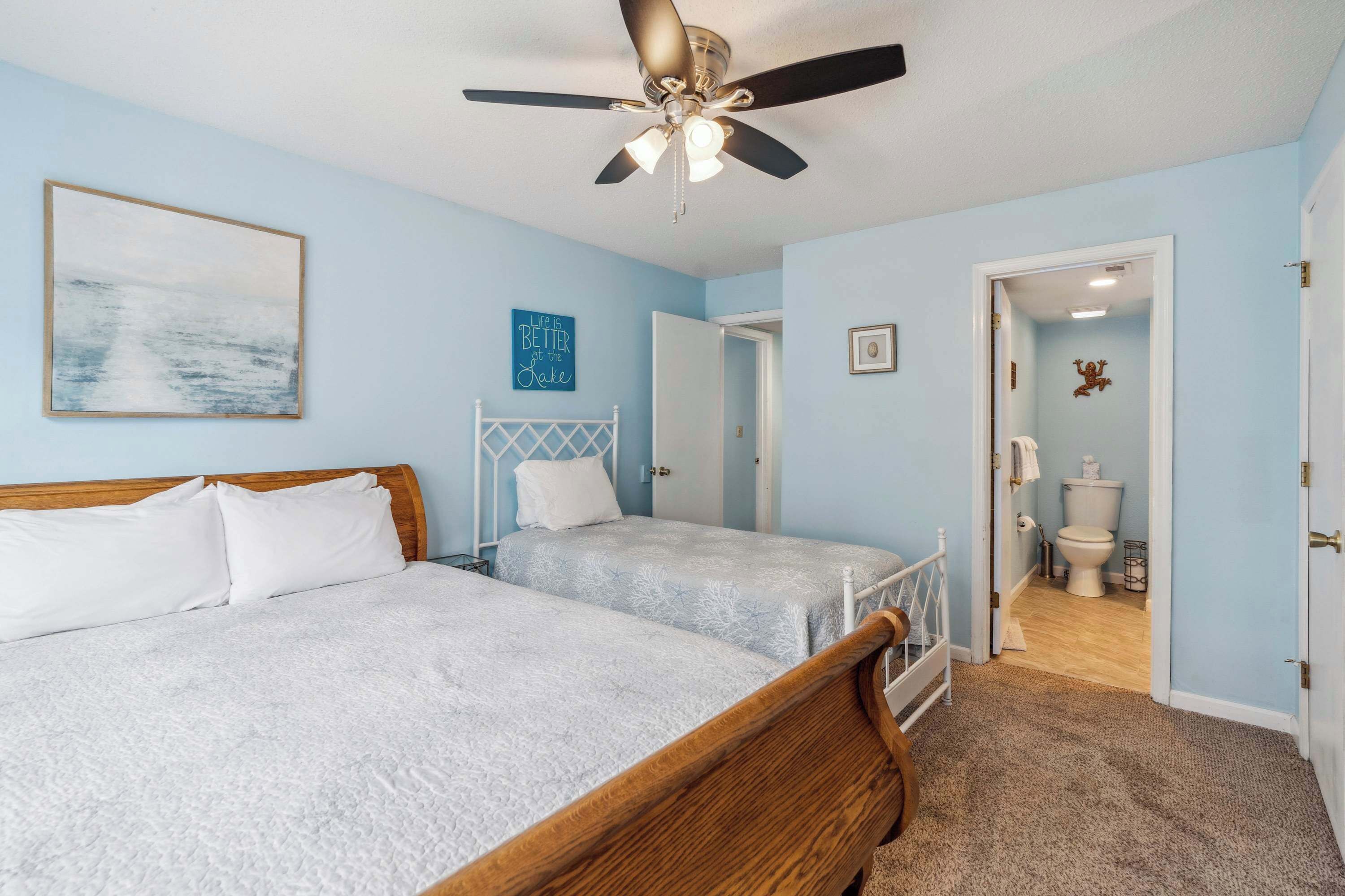 A bedroom with two beds, a ceiling fan, a painting, and a bathroom visible through an open door.
