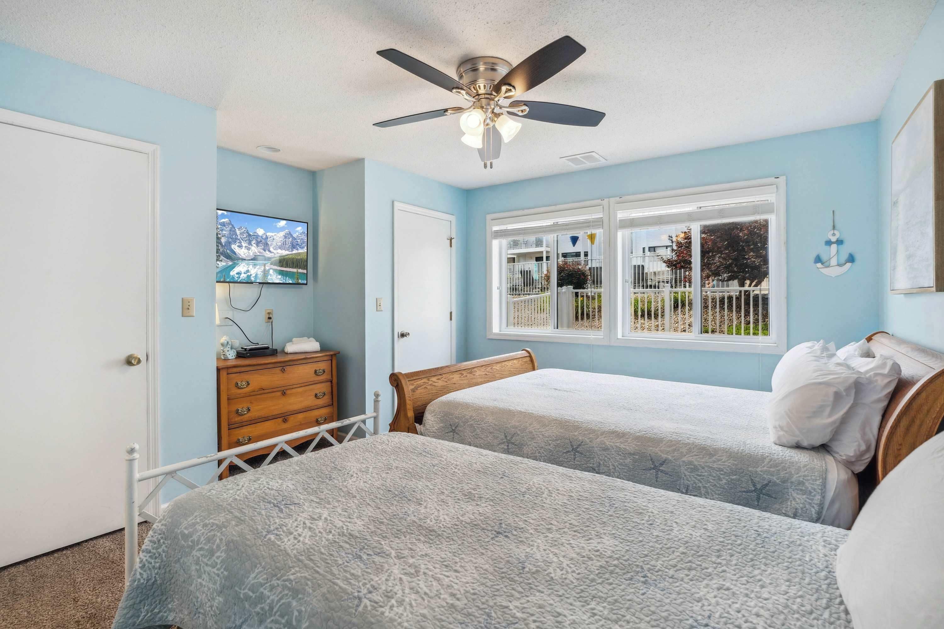 A bedroom with two beds, a ceiling fan, a TV, and a window with a view of a fenced area.