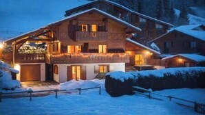 A wooden chalet with a balcony, snow-covered surroundings, and a lit entrance. - Simply Morzine - Apartment des Montagnes (Les Gets)