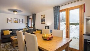 A living room with a sofa, chairs, a dining table with fruit, and a view of snow outside. - Simply Morzine - Apartment des Montagnes (Les Gets)