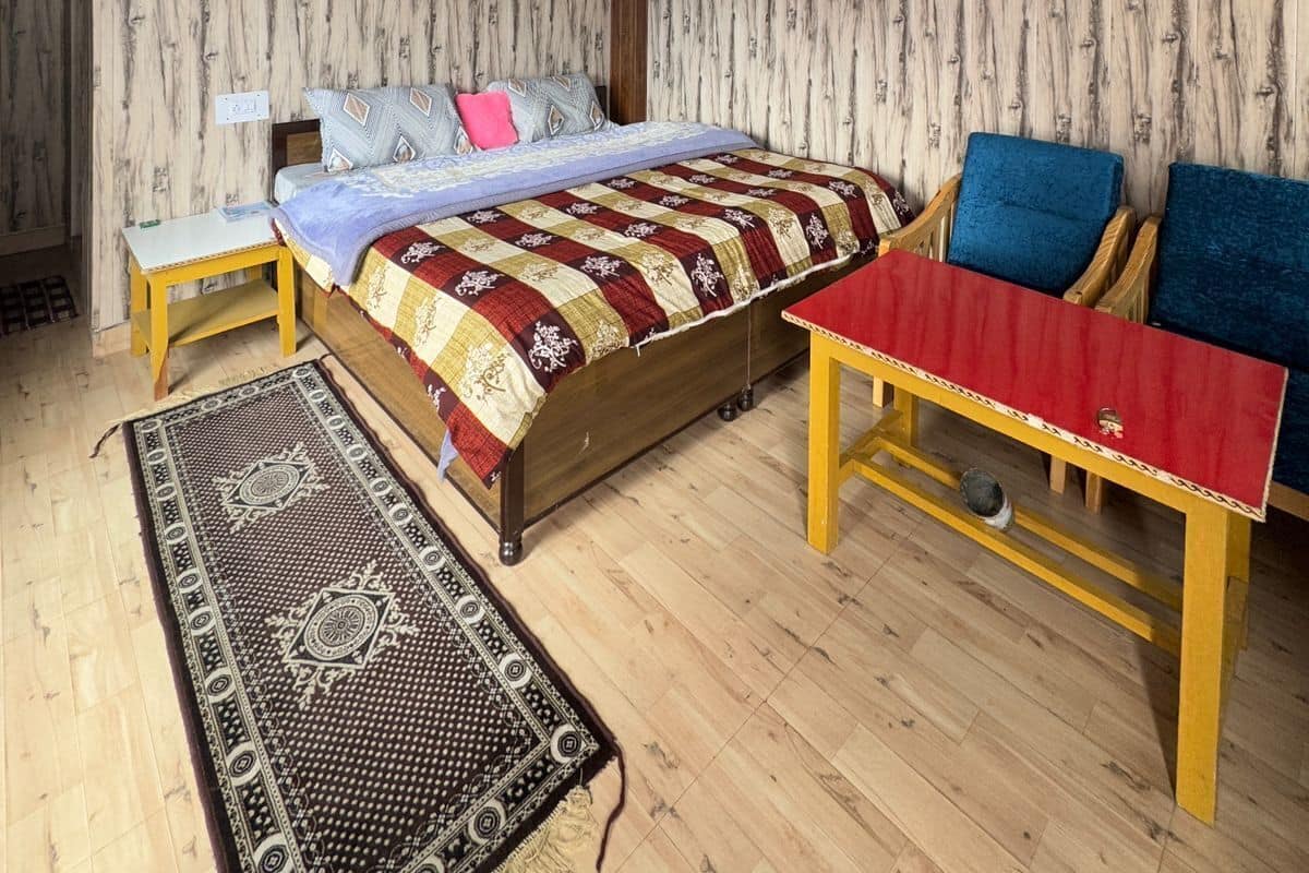 Une pièce avec un plafond en bois, un lit recouvert d’une courtepointe à motifs, une petite table avec une nappe rouge, deux chaises bleues et un tapis à motifs.