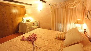 A bedroom with a large bed, a wooden wardrobe, a small table with a lamp, and a chair.