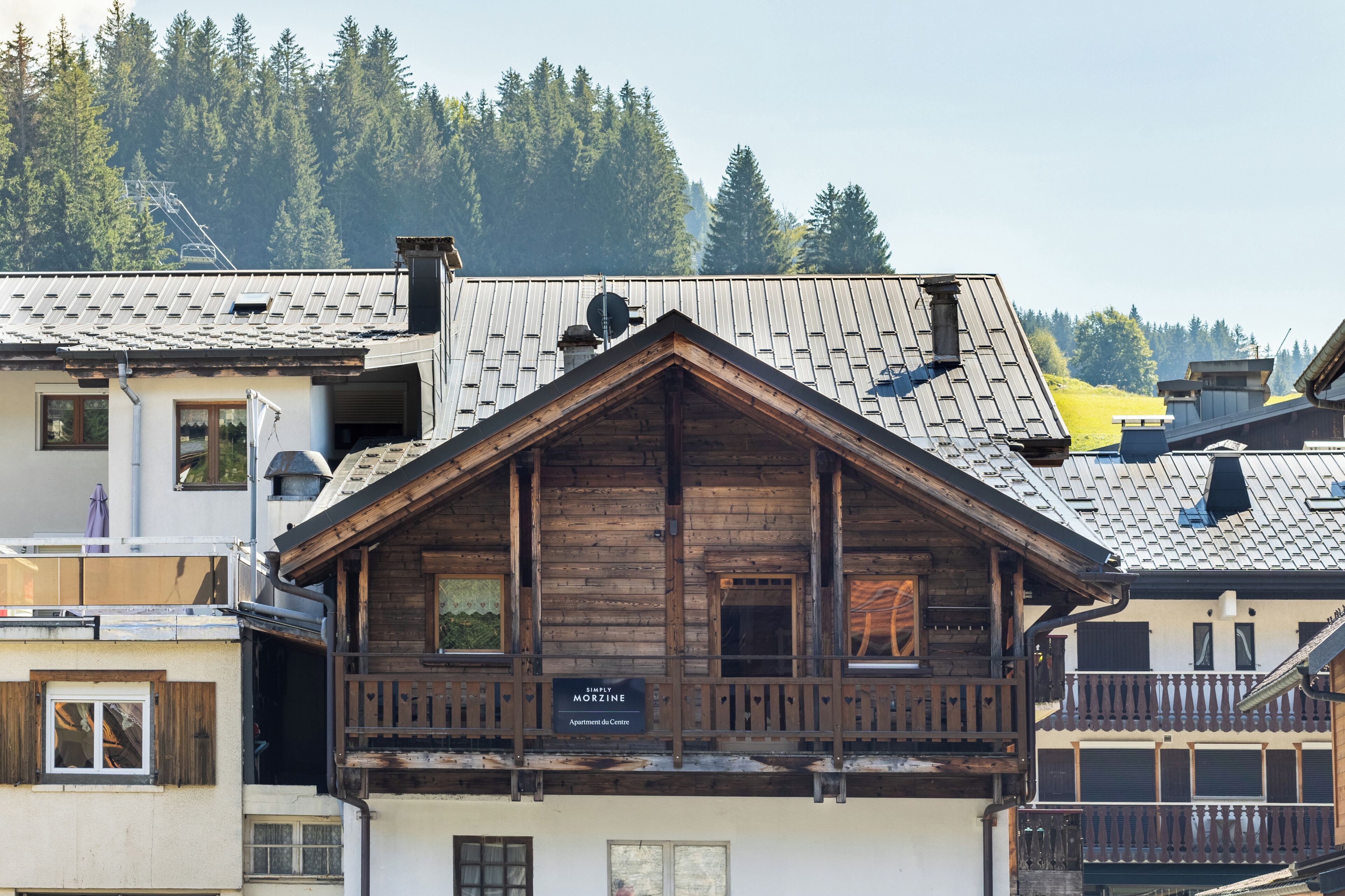 A wooden chalet with a balcony, a sign reading 'MORZINE Apartments de Centre,' and a mountainous backdrop.