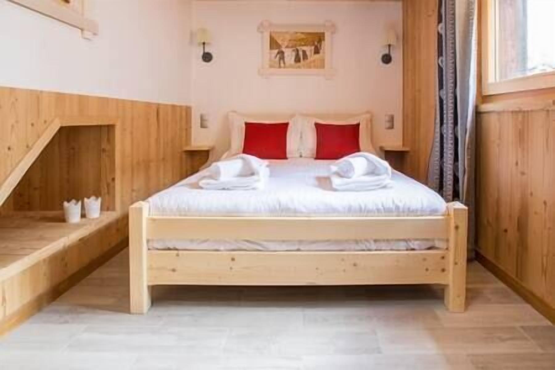 A wooden-framed bed with white bedding and red pillows, a small bedside table, and a wall-mounted light fixture.