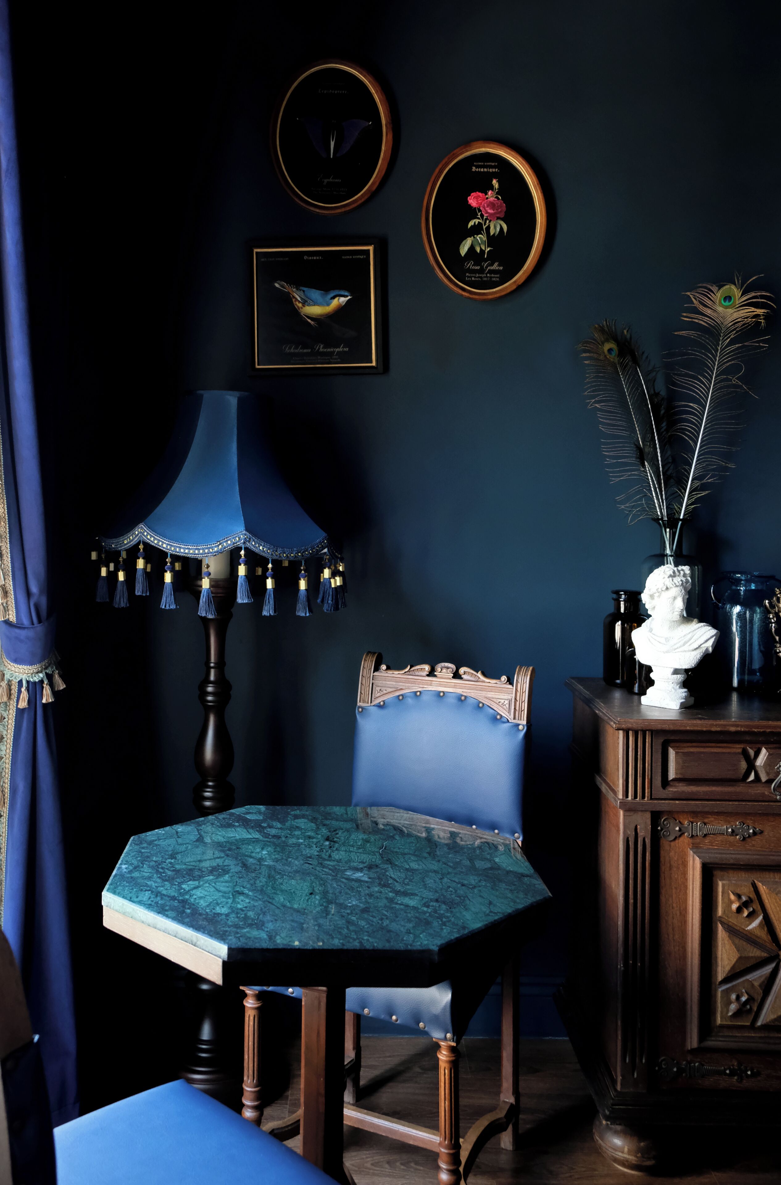 Una habitación con una mesa azul, una silla azul, una lámpara y adornos en la pared, incluyendo cuadros enmarcados y un busto.