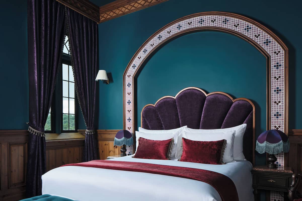 Quarto com cama grande, um banco, janela com cortinas e arco na parede revestida com azulejos.