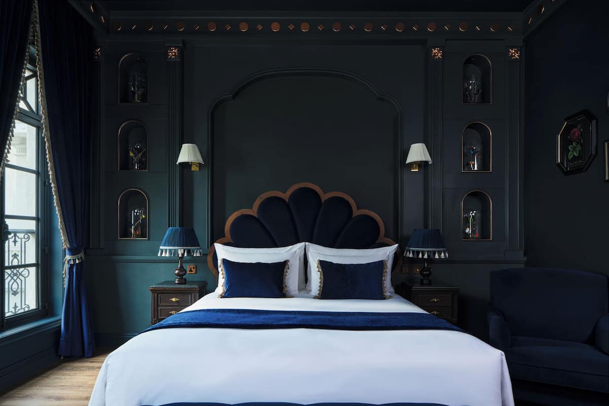 Quarto com uma cama grande, duas mesas de cabeceira com luminárias, parede escura com molduras decorativas e uma janela com cortinas.