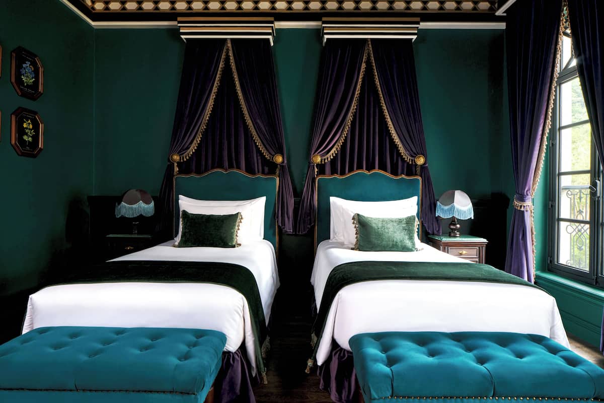 Quarto com duas camas, mesa de cabeceira, luminária e janela com cortinas.