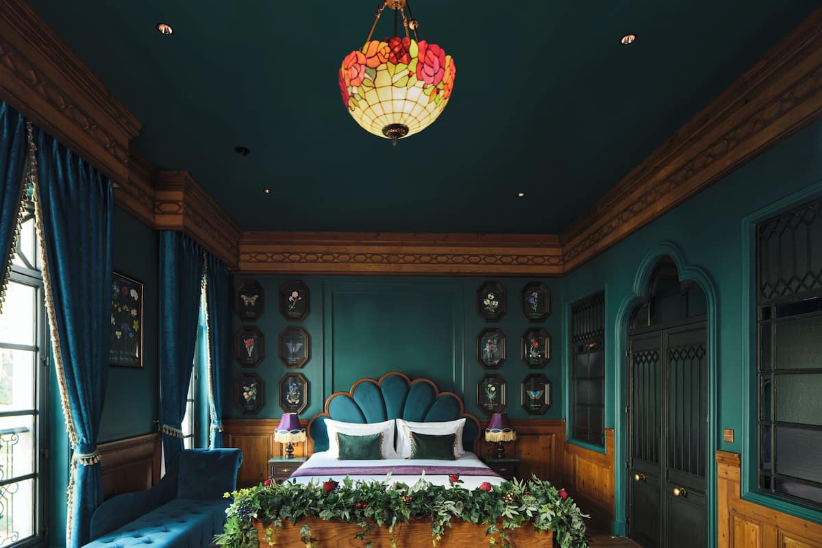 Quarto com cama grande, um banquinho, um candelabro e janela com cortinas.