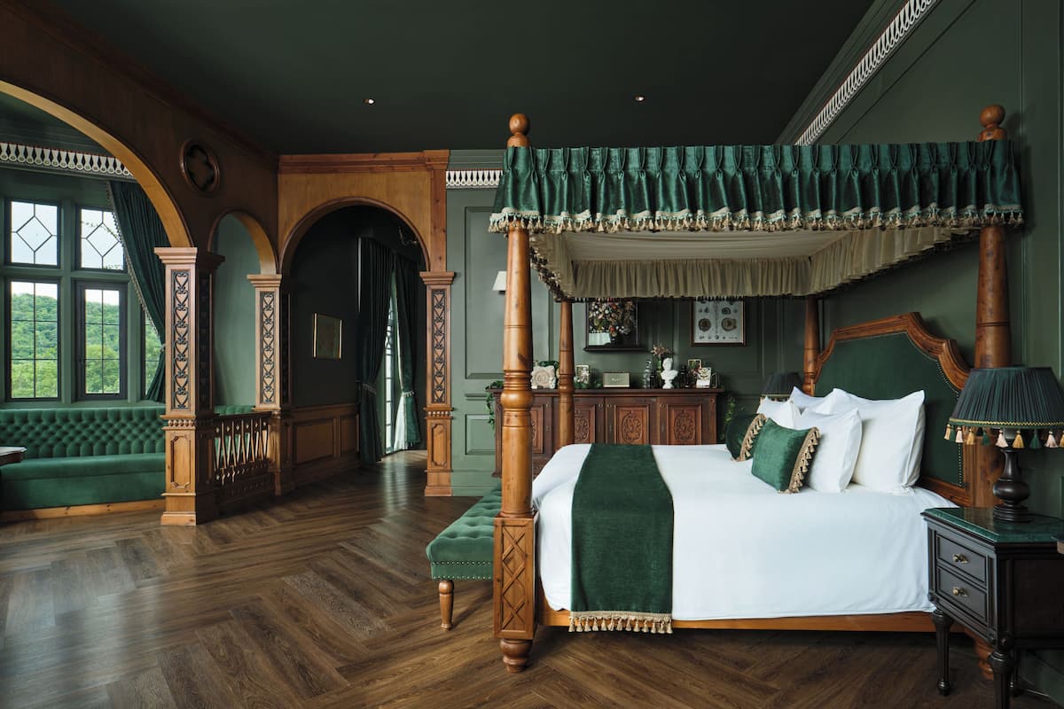 Uma cama de dossel com um edredom verde e almofadas combinando, um banquinho de madeira e uma mesinha de cabeceira com abajur.