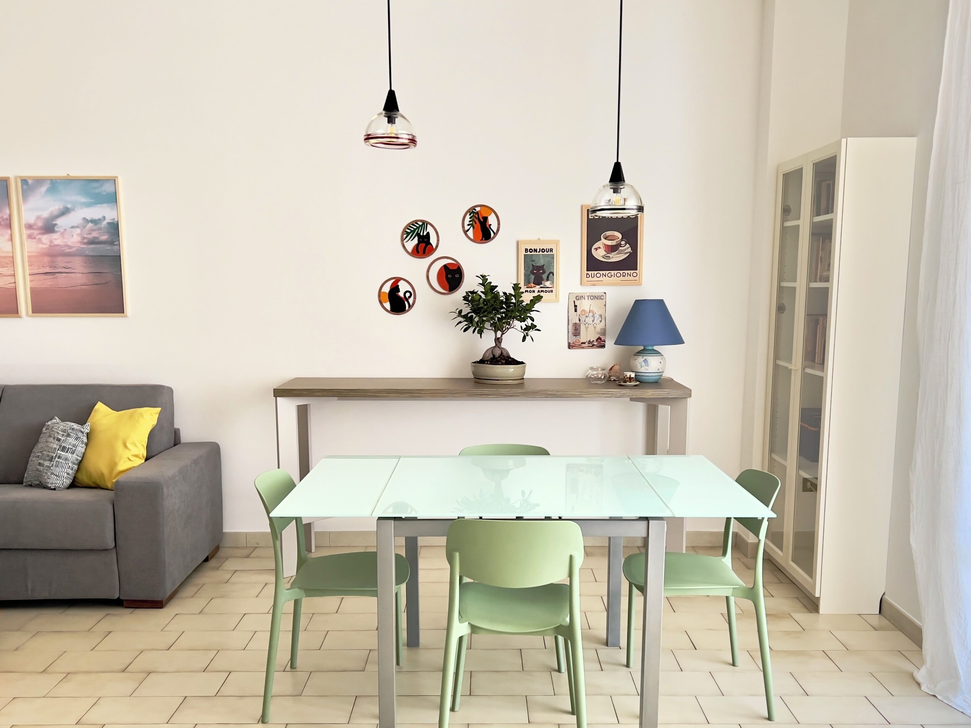 Un salon moderne avec un canapé gris, une table à manger en verre, des chaises vertes, une plante en pot et des œuvres d’art murales.